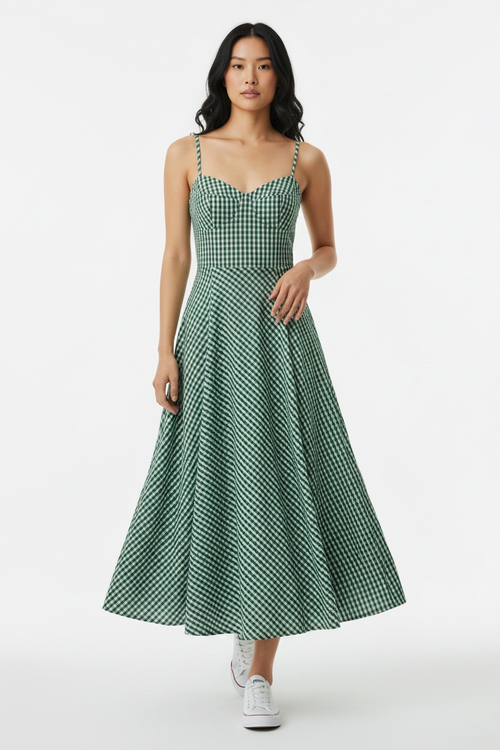 A woman modeling a green and white gingham midi dress with thin straps and a flared skirt, paired with white sneakers, against a plain light gray studio background.